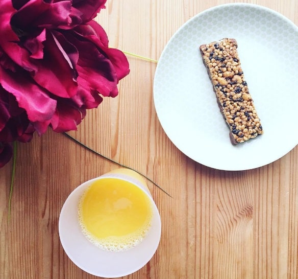 A cereal bar on a white plate, a glass of orange juice, and a large red flower sit on a wooden table—perfect for fuelling up before taking on the £2 Challenge by Voucherbox and Zamcog.