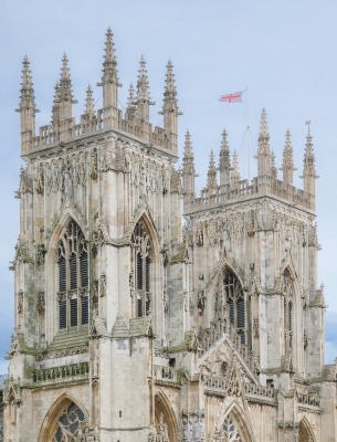 Gothic-style cathedral towers with detailed stone carvings and pointed arches, topped with spires. On a birthday trip to York, a flag with a red cross on white flies above one of the towers against a cloudy sky.