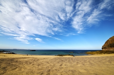 A sandy beach with calm blue sea waves under a bright sky filled with wispy clouds—an ideal spot for your first family holiday. On the right side, rocky cliffs rise above the horizon where the distant sea meets the sky.