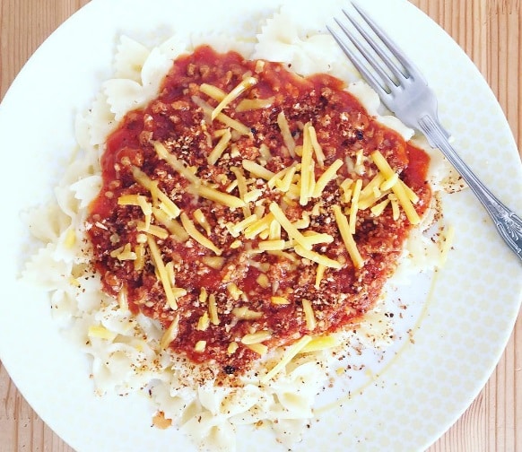 A plate of bowtie pasta topped with tomato sauce, grated cheese, and sprinkled herbs sits on a light wooden surface—perfect for a £2 Challenge meal inspired by Zamcog and Voucherbox. A fork rests at the plate’s edge, ready to enjoy.