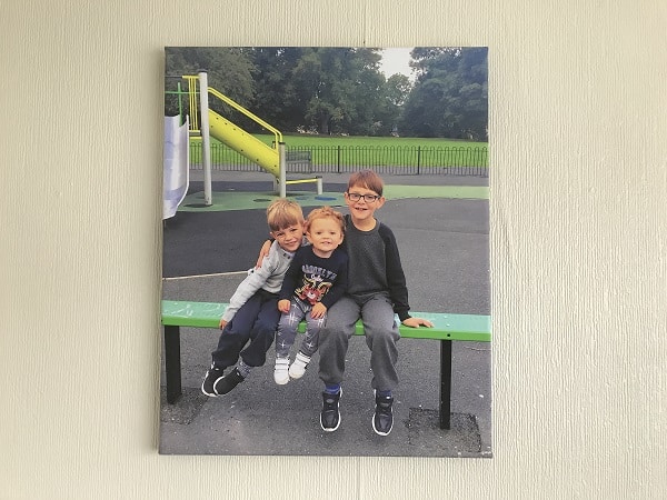 A photo of three young children sitting close together on a green bench at a playground, smiling at the camera. Perfect to display photos of your favourite moments around the house, it features a yellow slide and green grass in the background.
