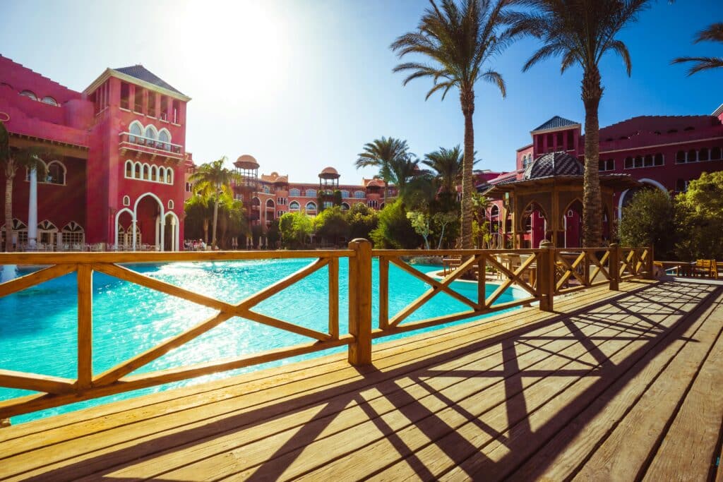 A sunny resort scene with a large, clear blue swimming pool, palm trees, and red buildings in the background—perfect for your travel bucket list. Wooden railing and decking cast long shadows across the foreground.
