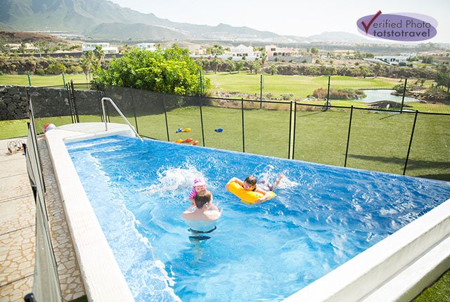 Three children play in a small outdoor pool on a sunny day, surrounded by grass and a fence—capturing pure joy on their first family holiday, with rolling hills, houses, and mountains stretching into the background.