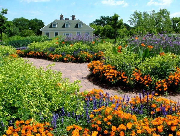 A vibrant garden design with curved brick pathways, colourful flowers in orange, yellow, and purple, and lush green bushes. In the background, a house with dormer windows is partially visible under a blue sky.