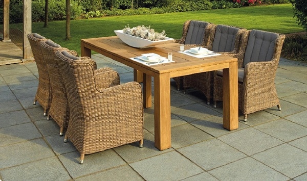 A wooden outdoor dining table with six wicker chairs sits on a tiled patio, perfect for garden style entertaining. The table is set with plates, cutlery, and a white centrepiece. Green grass and plants create a lush backdrop.
