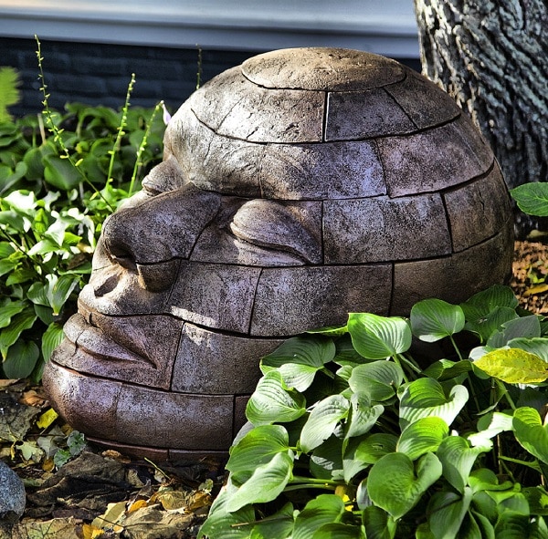A large stone sculpture of a human face, composed of block-like segments, rests on the ground surrounded by lush green plants and leaves, creating a striking focal point for inspired garden design.