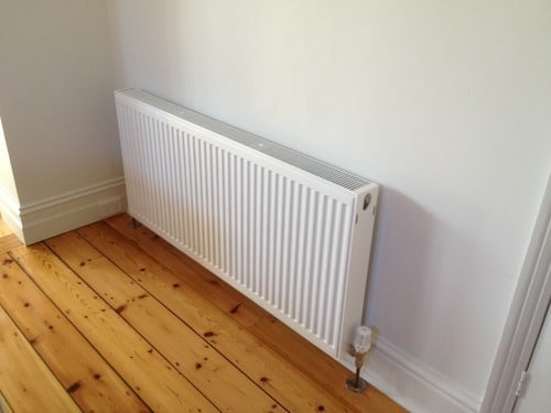 A white metal radiator attached to a white wall, positioned above light-coloured wooden floorboards in a corner, shows how to heat your home efficiently while blending seamlessly with modern décor.