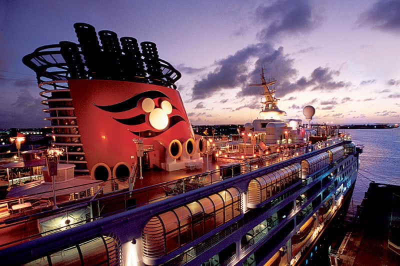 A Disney Cruise Line ship at sunset, featuring illuminated decks and a large red funnel with Mickey Mouse ears and logo, promises the perfect family holiday cruise, berthed near calm water under a partly cloudy sky.