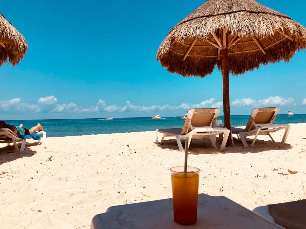 A tropical beach scene perfect for a beach holiday with your family: straw parasols, empty sun loungers on white sand, a drink on a table in the foreground, clear blue sky, and boats anchored in the calm sea water beyond.