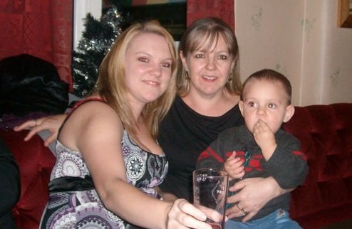 Two women sit closely together on a red sofa, smiling at the camera, while a young boy sits on Mum’s lap, eating with his hand. A Christmas tree glows in the background, capturing memories from their festive journeys together.