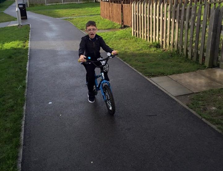 A young boy wearing glasses and dark clothes rides a blue bicycle on a paved path in a park, enjoying one of many outdoor activities for children, with grass and a wooden fence alongside the path.