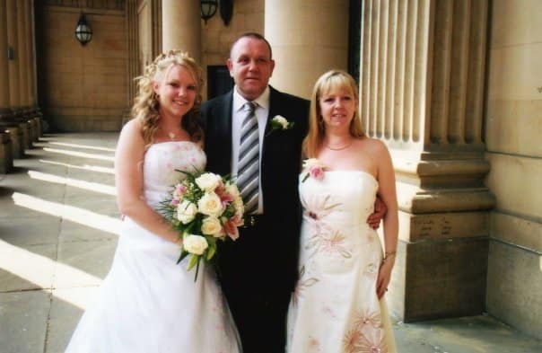 Three people stand together in formal attire outside a columned building. The man in the centre wears a suit and tie, while the women beside him, in white dresses holding bouquets, create a scene reminiscent of "Journeys With My Mum" at a wedding.