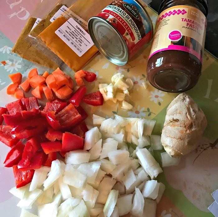 Chopped onions, red pepper, carrots, and a piece of ginger are on a chopping board—perfect for cooking healthy meals with curry powder sachets, tomato purée, and tamarind paste from the WW Smart Kitchen collection nearby.