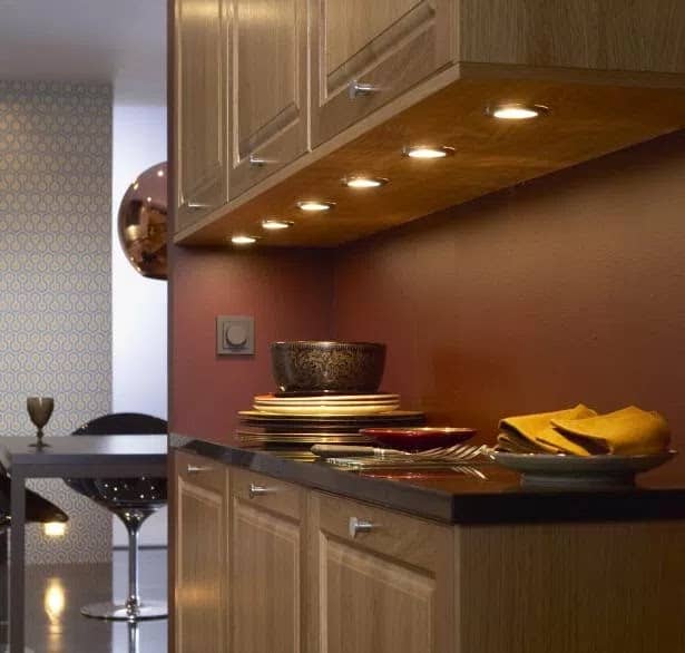 A modern kitchen features wooden cupboards with under-cupboard LED lighting. Plates, bowls, and folded yellow serviettes rest on a dark worktop. A patterned wall and a bar stool are visible in the background.