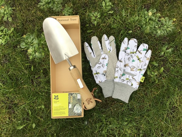A gardening set on grass, featuring a metal trowel with a wooden handle in a box, patterned gardening gloves with leaf and bird designs, and a National Trust info card—an ideal Mother’s Day gift featured on the 2017 Gift List.