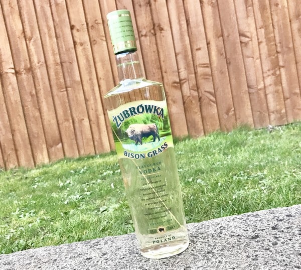 A bottle of Żubrówka Bison Grass Vodka stands on a stone surface outdoors, with green grass and a wooden fence in the background. The bottle, ideal for any Gift List or Mother’s Day 2017 celebration, has a green cap and a label featuring a bison.