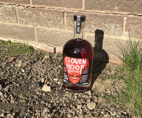 A bottle of Cloven Hoof Spiced Rum sits on gravel near a brick wall, sunlight casting a shadow and some green grass to the right—an ideal addition to your 2017 Mother’s Day Gift List.