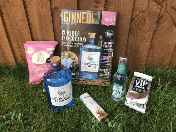 A selection of items on grass in front of a wooden fence: Gunpowder Irish gin bottle, tonic water, GINNED! magazine, pink crisps packet, snack bar, and a pack of wasabi peas—perfect for a 2017 Gift List or Mother's Day treat.