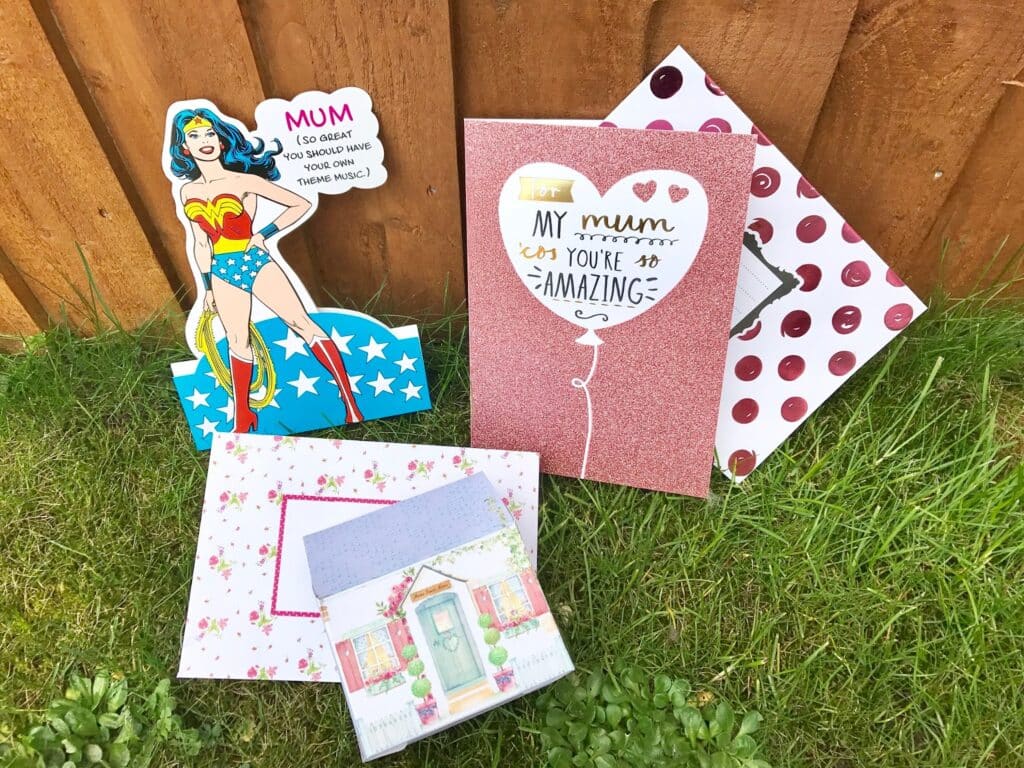 A collection of colourful 2017 Mother’s Day cards, including a Wonder Woman themed card, laid out on green grass in front of a wooden fence—perfect inspiration for your Mother’s Day gifts or gift list.