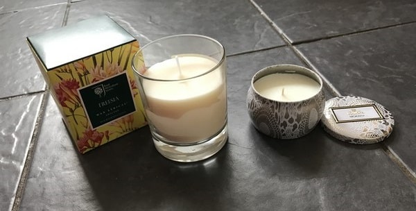 Three candles on dark tiles: a glass holder with a yellow floral box, perfect for a 2017 Mother’s Day Gift List; a silver tin with a decorative lid beside it; and a small white container with matching lid.