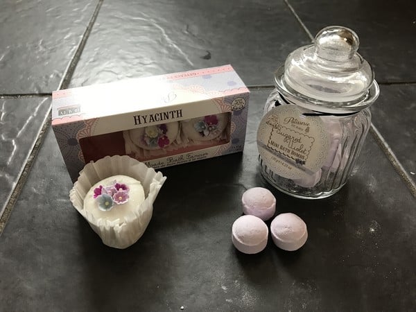 A box of floral-decorated hyacinth-scented bath fizzers, a glass jar labelled and lidded with purple bath bombs, and three small round purple bath bombs on a dark tiled surface—perfect for any Mother’s Day gifts list.