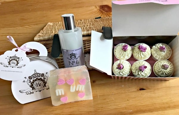 A wooden table displays a box of six cupcakes topped with white icing and pink flowers, a bottle of lotion, a bar of soap with pink hearts, and decorative tags—perfect for your 2017 Mother’s Day gift list.