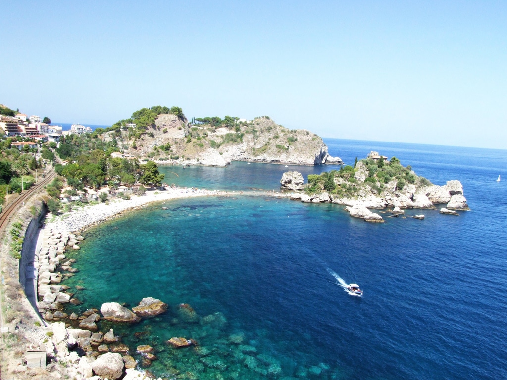 Aerial view of a scenic Sicilian coastal bay with clear blue water, rocky islands, and lush greenery. A small boat moves across the bay, while the curved shoreline and nearby railway track promise a perfect Sicilian family adventure.