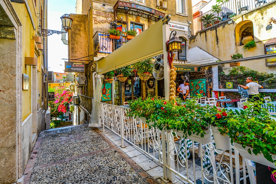 A colourful, narrow alleyway in a Sicilian town features potted plants, a cobblestone path, outdoor café seating, and vibrant façades. Ideal for Sicily lovers seeking family travel tips or picturing their next Sicilian family adventure.