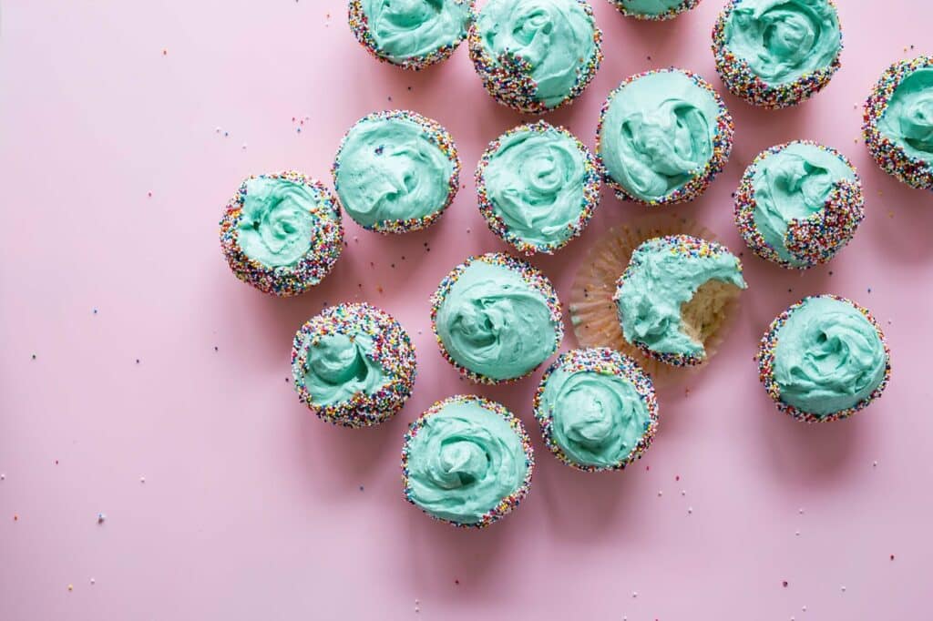 Cupcakes with light blue icing and colourful sprinkles are arranged on a pink surface. One cupcake has a bite taken out of it, offering sweet inspiration for anyone wanting to start a baking business.