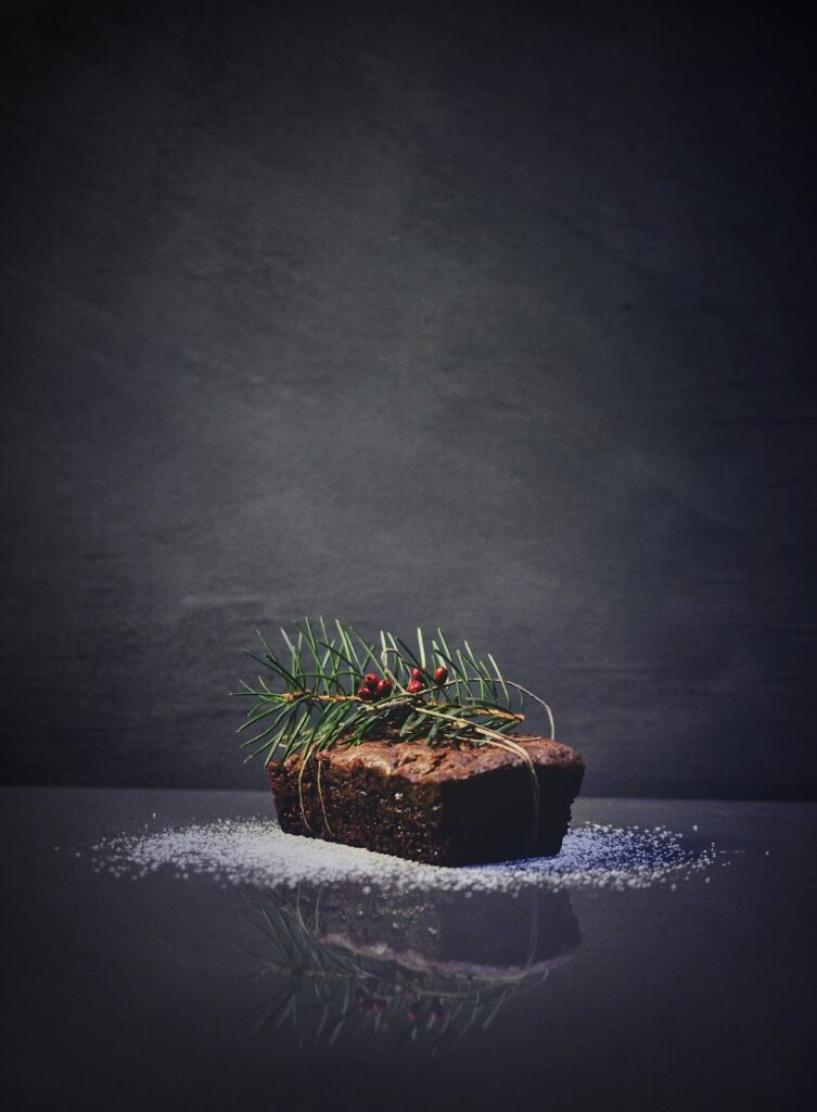 A small chocolate loaf cake tied with string, topped with pine needles and red berries, sits on a dark reflective surface dusted with icing sugar—perfect inspiration if you want to start a baking business or need fresh holiday baking tips.