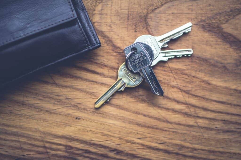 A set of three silver keys on a keyring rests on a wooden surface next to a dark leather wallet, evoking the excitement of buying a house and stepping into your new home.
