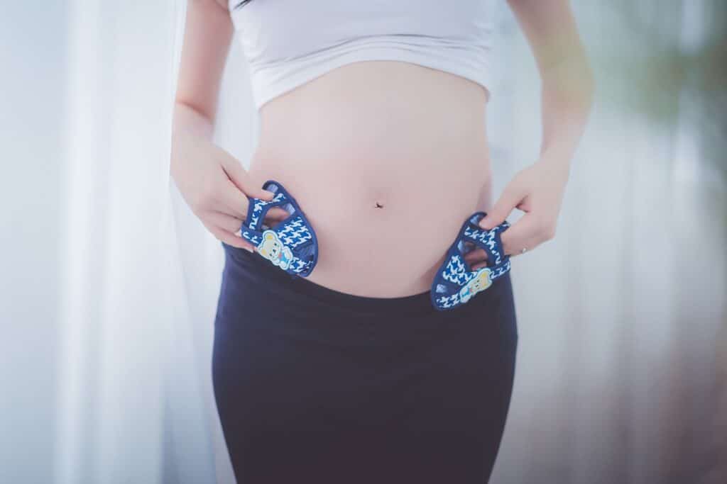 A pregnant person wearing a white top and black skirt holds a pair of small blue baby shoes against their bare tummy, symbolising anticipation of a baby and the journey of fertility, wondering what to expect in the months ahead.
