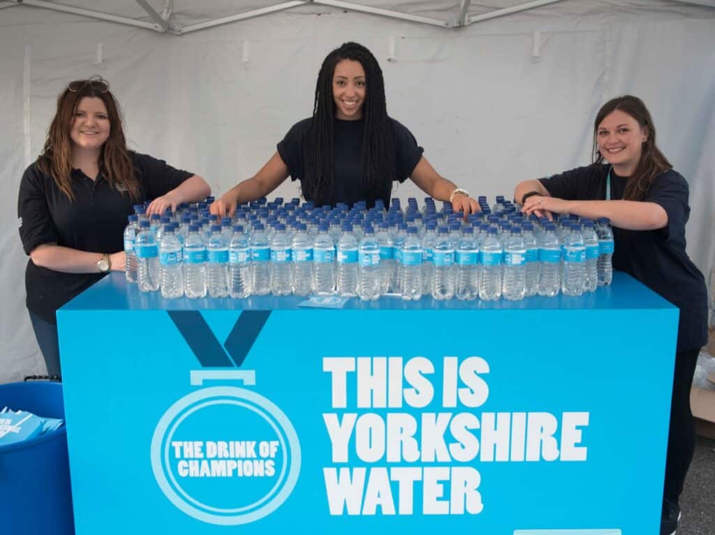 Three smiling working women stand behind a table covered with Yorkshire Water bottles. The table displays a blue sign reading, "THIS IS YORKSHIRE WATER" and "THE DRINK OF CHAMPIONS," complete with a medal graphic, under a white marquee.