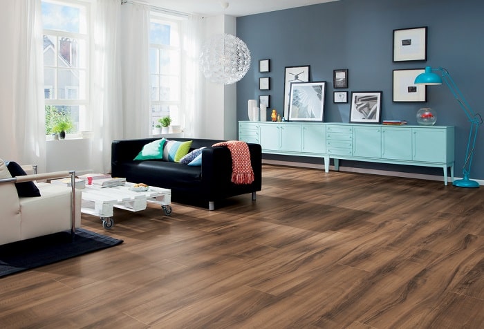 Bright, modern living room with laminate wood flooring, a black sofa, white coffee table on castors, turquoise sideboard, blue standard lamp, framed art on a grey wall, and large windows with white curtains letting in natural light.