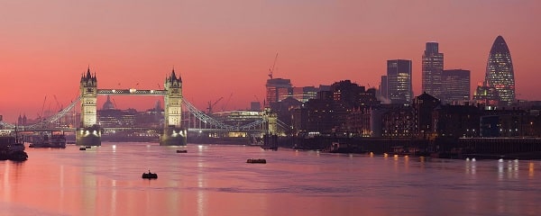 Tower Bridge and the London skyline at sunset, with the River Thames reflecting the sky’s pink and orange hues. Several boats glide by—a perfect view after a day of exploring family attractions London has to offer.