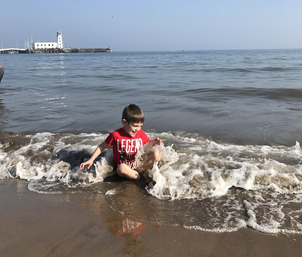 A young boy wearing a red "LEGEND" shirt sits in shallow seaside waves at the edge of a sandy beach, smiling and playing as water splashes around him. A pier and building are visible in the background under a clear blue sky.