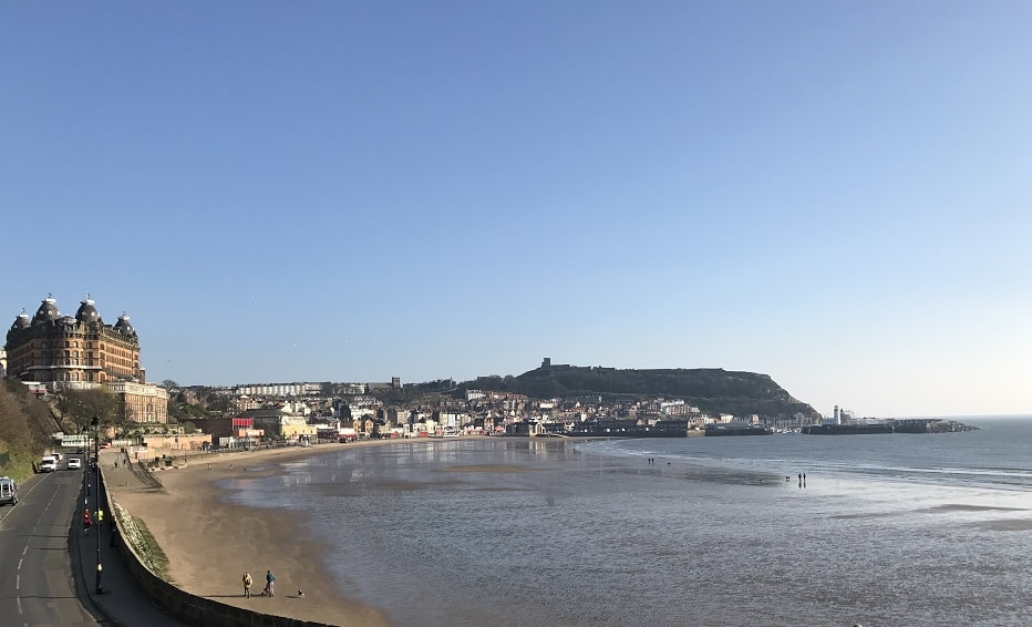 A wide sandy beach curves along a Yorkshire seaside town with historic buildings on the left, a castle-topped hill in the distance, and a calm blue sky overhead. People walk along the shore and a sea wall lines the road, capturing classic countryside charm.