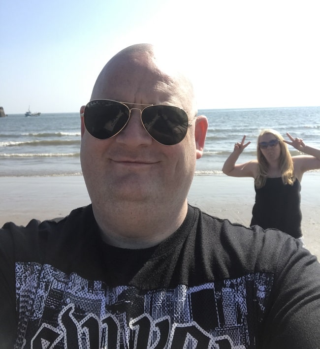 A man wearing sunglasses takes a selfie on a sunny beach, smiling at the camera. In the background, a woman—perhaps taking a break from relationship issues—poses playfully with peace signs near the shoreline as gentle waves roll in.