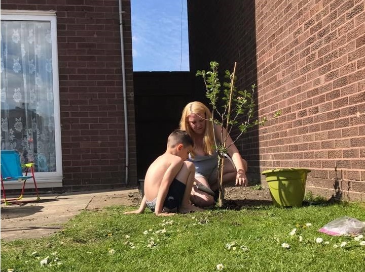 A woman and a young boy are planting a small tree next to a brick building on a sunny day. Engaged in kids' gardening, they focus on the task as gardening tools and a green pot sit nearby.