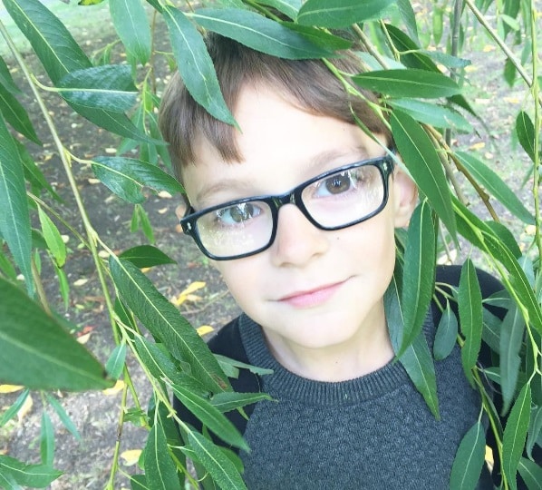 A young boy with short brown hair and glasses looks up through green leafy branches in a children’s garden, wearing a dark grey jumper, with sunlight and patches of grass visible in the background.