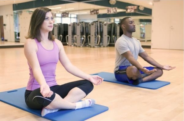Two people sit cross-legged on yoga mats in a gym studio, practising post-pregnancy exercise or meditation with eyes open and hands on knees. Mirrors and gym equipment in the background create an ideal space for weight loss after pregnancy.