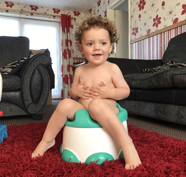 A curly-haired toddler sits on a potty in a living room, smiling with hands on their chest—capturing a sweet potty training moment amid patterned wallpaper, a red rug, dark settees, and sunlight streaming through the window.