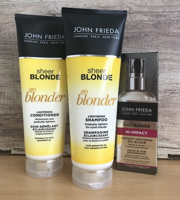 Three John Frieda Sheer Blonde hair products are displayed: a lightening conditioner, a lightening shampoo, and a hi-impact vibrancy restoring oil elixir from the Blonde Collection, all arranged on a wooden surface.