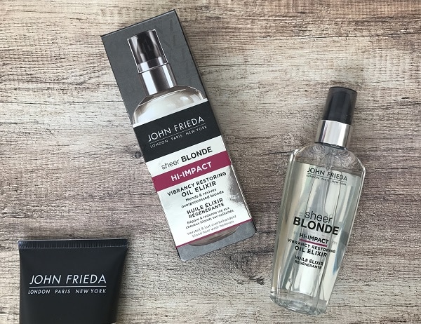 A bottle of John Frieda Sheer Blonde Hi-Impact Oil Elixir, its packaging box, and a black tube from the John Frieda Blonde Collection are arranged on a wooden surface.