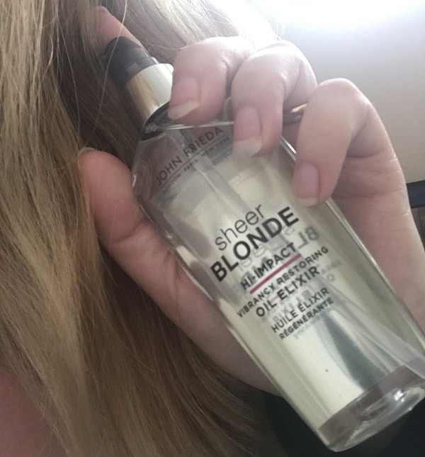 A hand holds a bottle of John Frieda Sheer Blonde Hi-Impact Oil Elixir near long blonde hair, highlighting its use for enhancing and nourishing blonde hair.