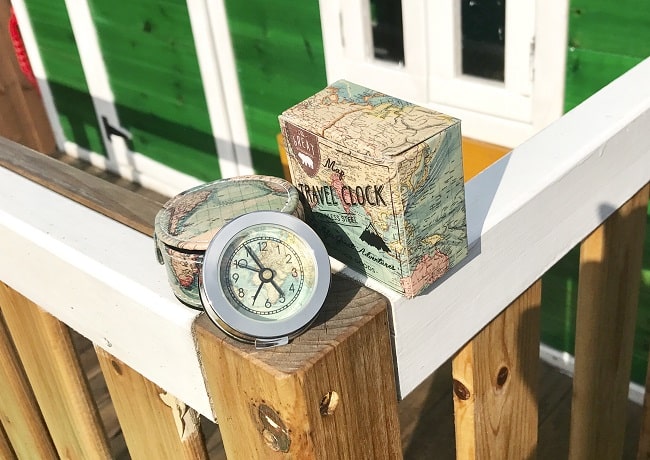 A small round travel clock in a map-patterned case sits on a wooden railing next to its matching box—a perfect quirky gift from Qwerkity. A green building with white trim is visible in the background.