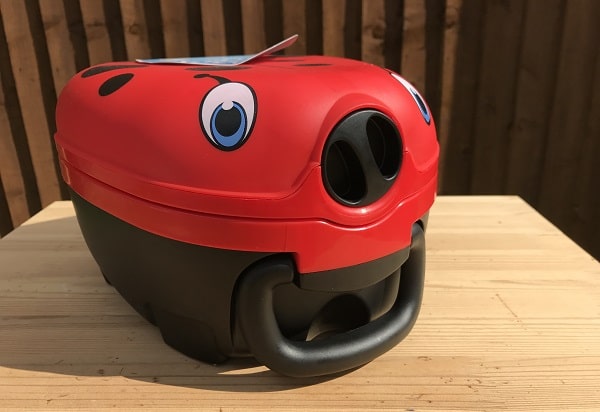 A red and black vacuum cleaner designed to look like a cartoon ladybird with large eyes, perfect for toddler spaces or potty training areas, sits on a wooden table in front of a wooden fence.