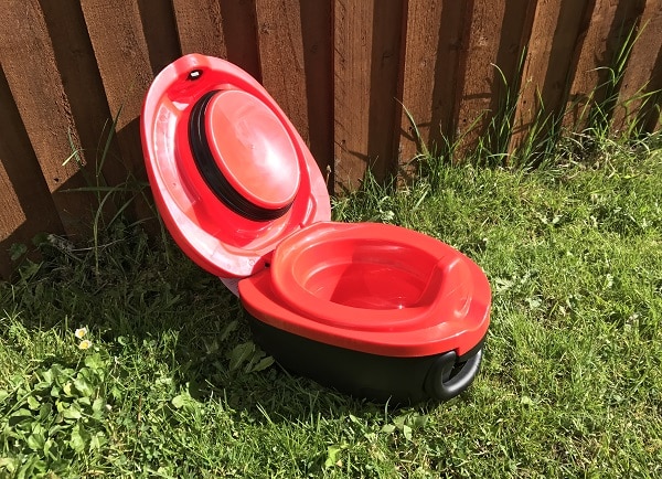 A red and black portable camping toilet, ideal for potty training a toddler, sits on green grass next to a wooden fence, its lid open and ready for use.