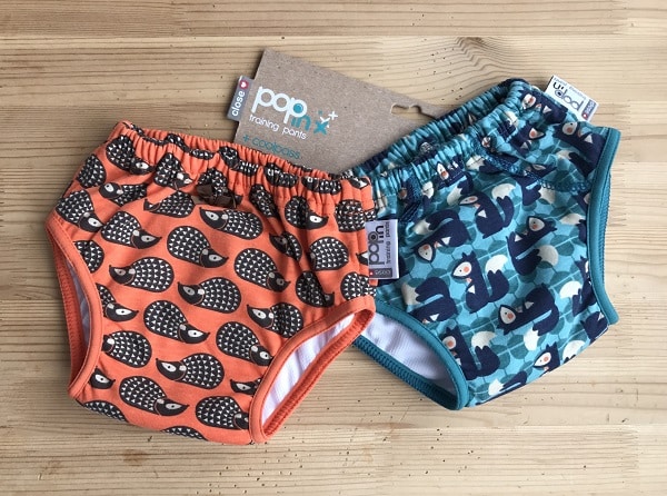 Two pairs of toddler training pants on a wooden surface, perfect for potty training. One pair is orange with a hedgehog pattern, the other blue with bears. Both feature elastic waistbands and attached product tags.