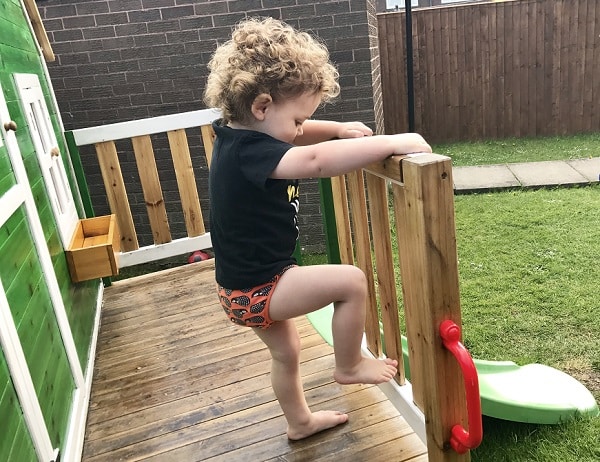 A toddler with curly hair, wearing a black shirt and patterned shorts, climbs over the wooden railing of a playhouse onto a grassy garden—taking adventurous steps even during potty training.